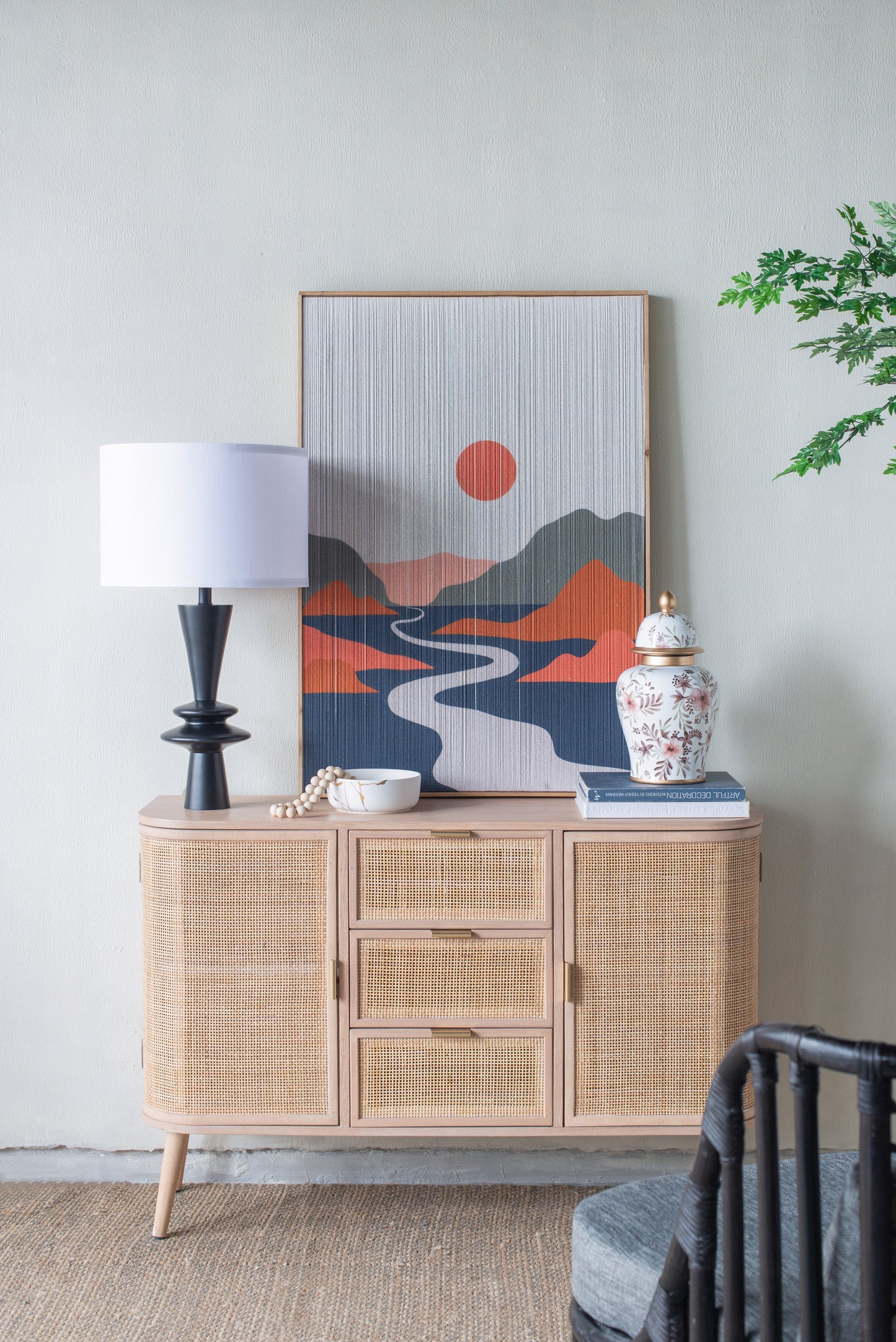 Black lamp with a white drum shade on a wicker sideboard with decorative items and a mountain painting in a room.