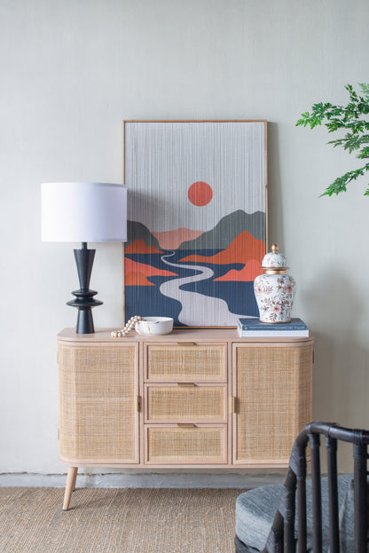 Black lamp with a white drum shade on a wicker sideboard with decorative items and a mountain painting in a room.