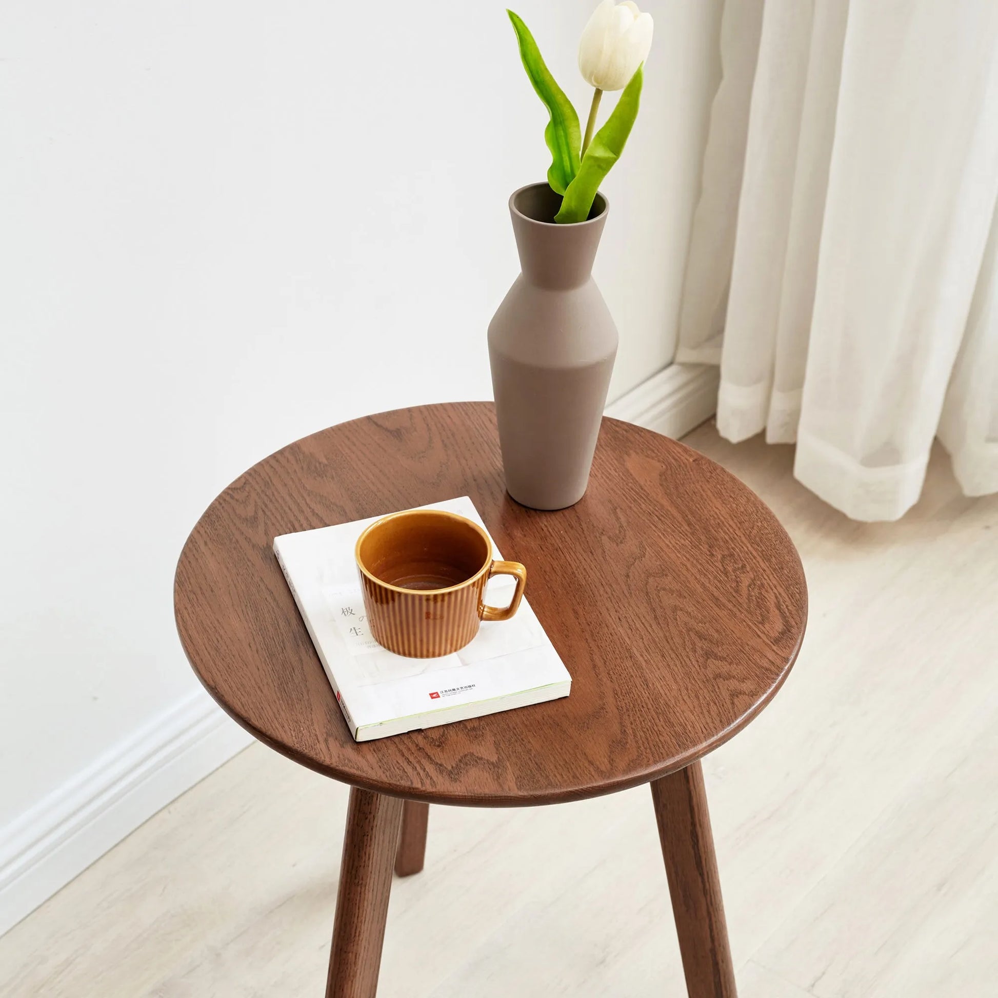 Small wooden end table with a cup, book, and vase against a white curtain background