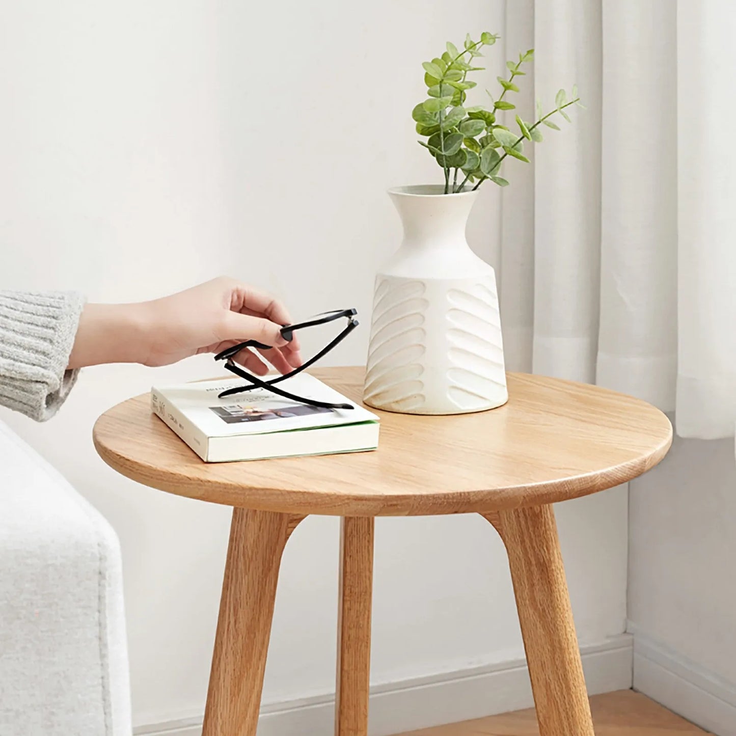 Round wooden end table with a white vase and green plant in a minimalistic room.