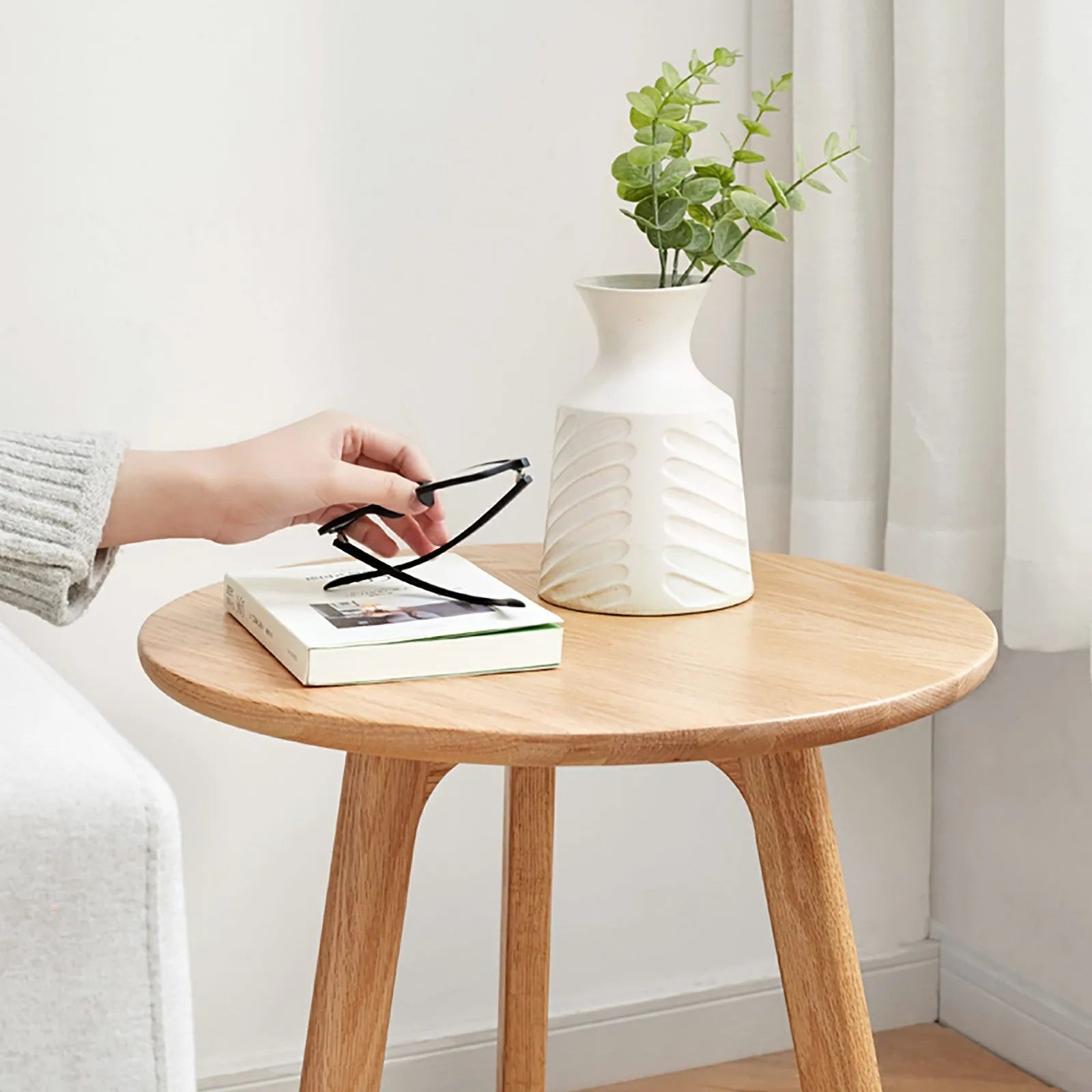 Round wooden end table with a white vase and green plant in a minimalistic room.