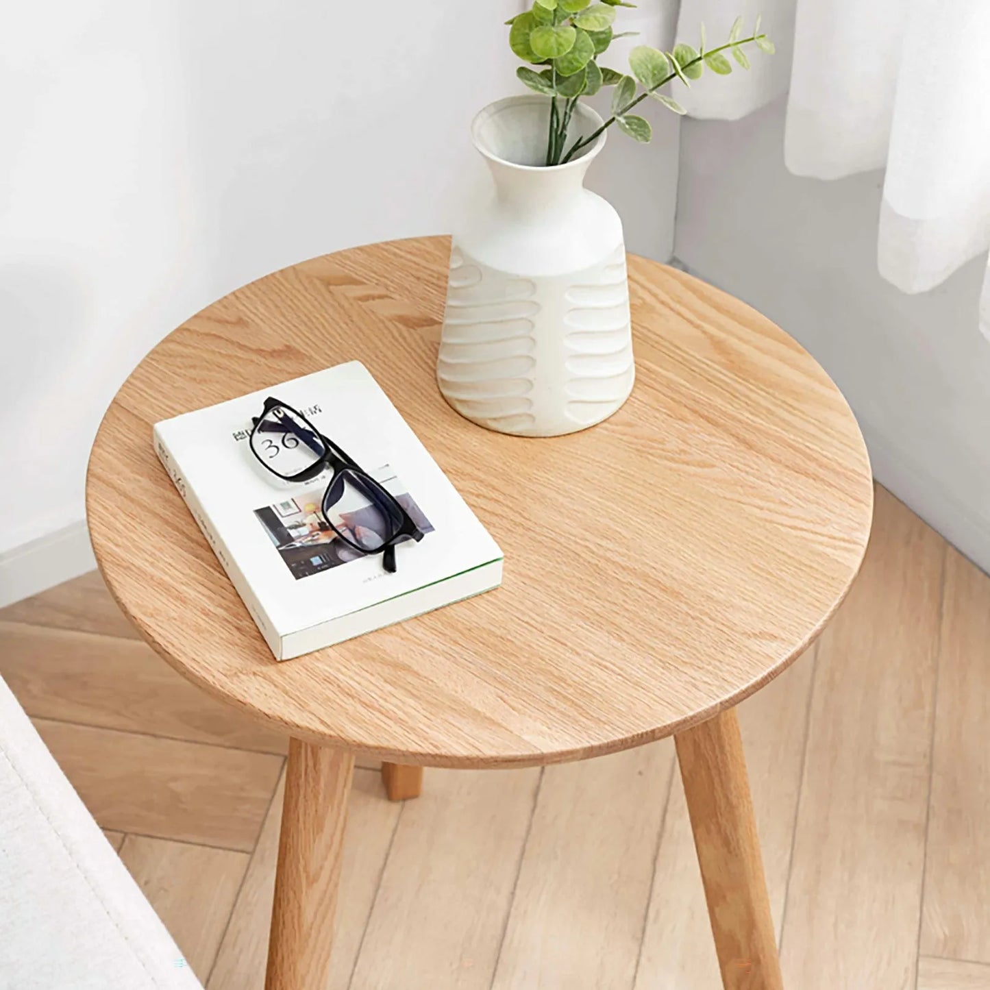 Round wooden end table with a book, glasses, and a vase on a light wood floor.