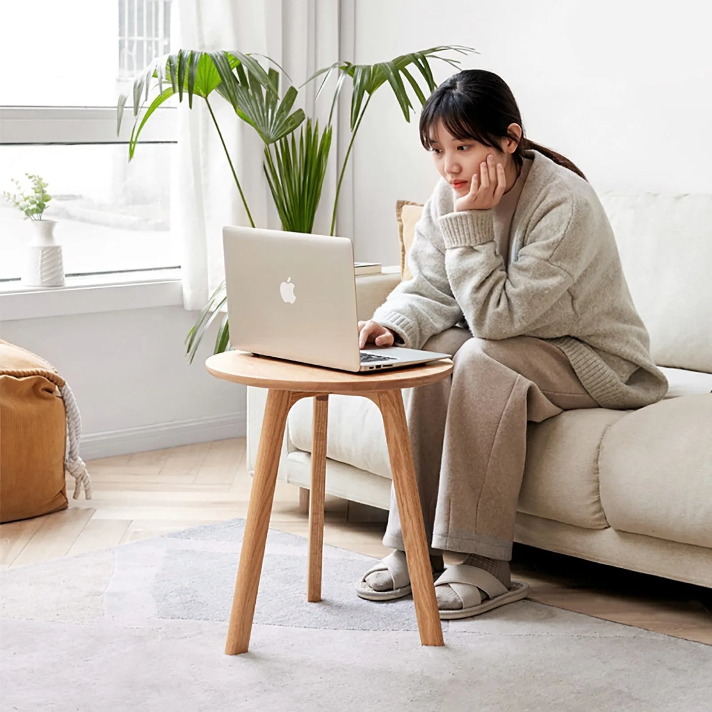 Woman using a laptop on a small wooden accent table in a bright living room.