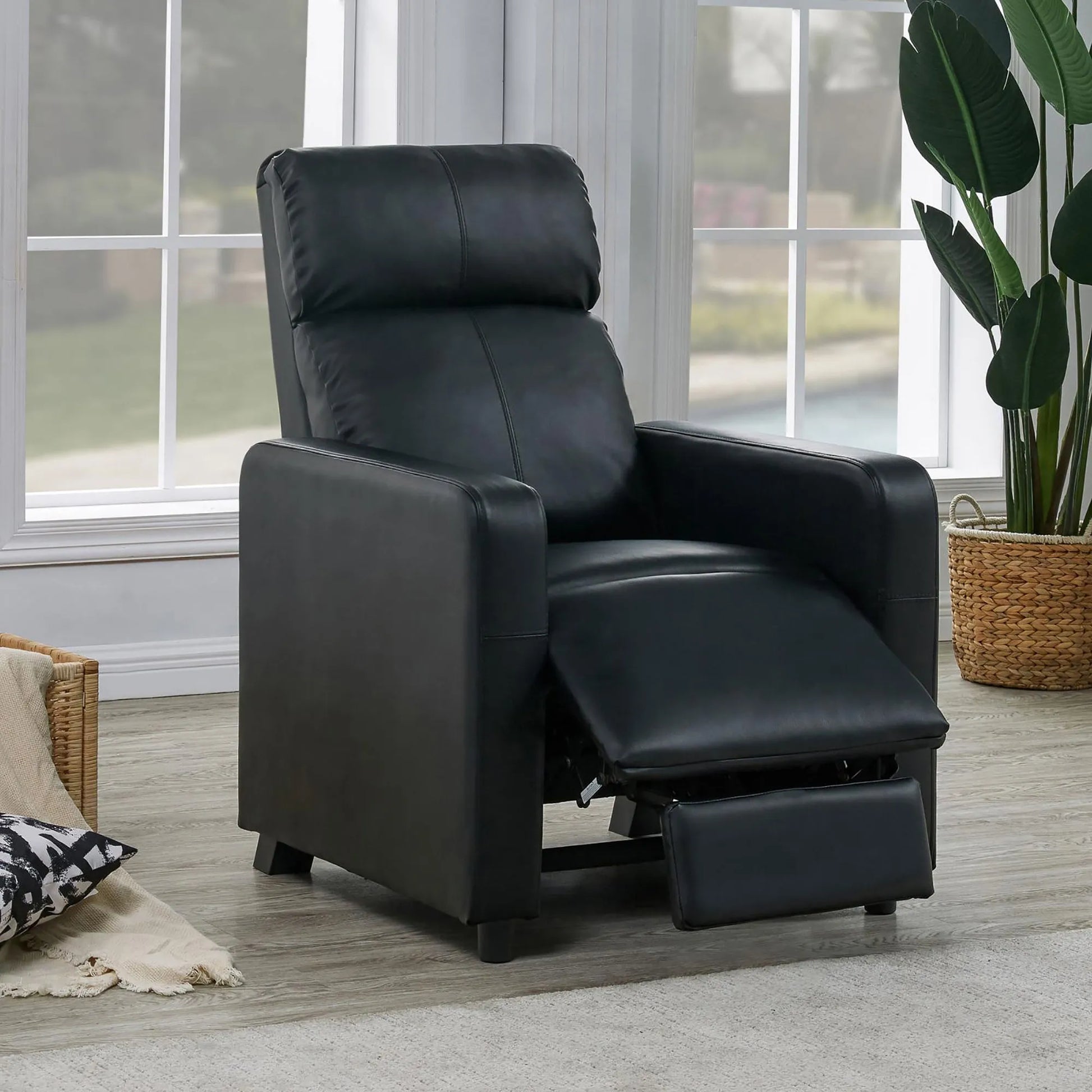 Black leather recliner chair with footrest extended in a living room setting with windows and a plant.