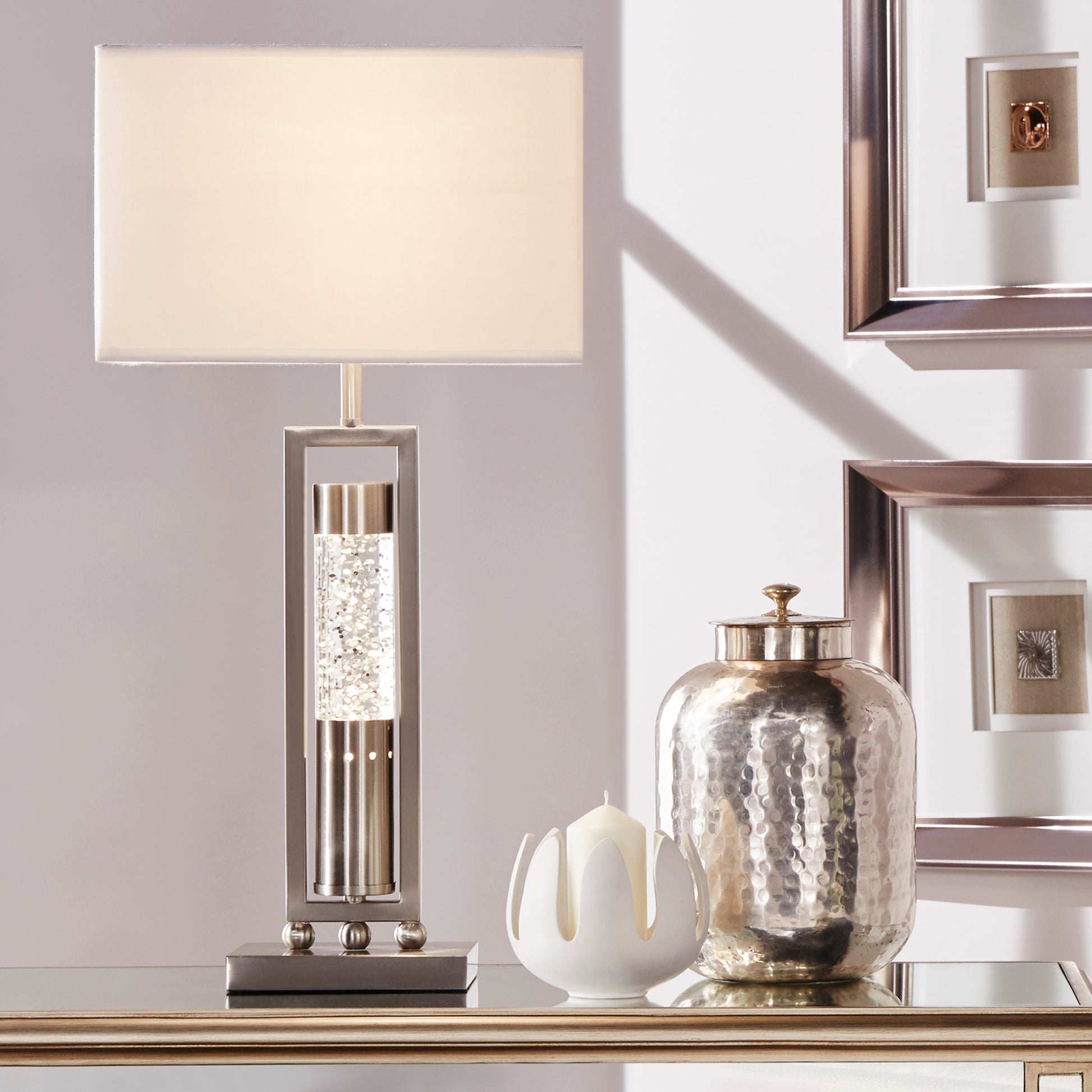 Decorative table lamp with a textured base and white shade on a surface with a mirror and candle in the background.