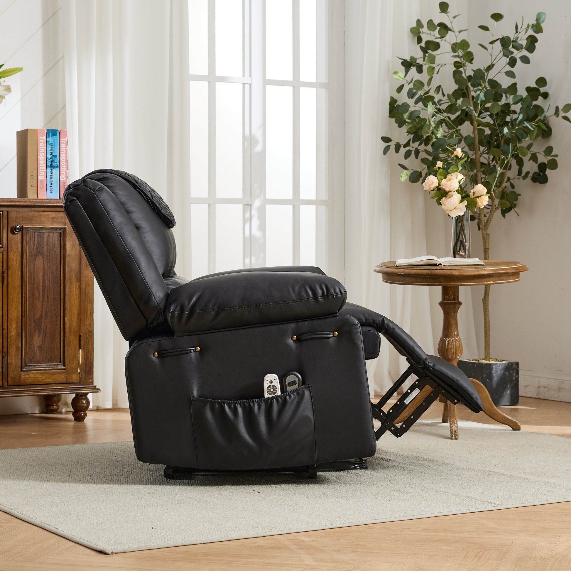 Side view of a black recliner chair with storage pocket and footrest extended in a living room setting with a wooden cabinet and table in the background.