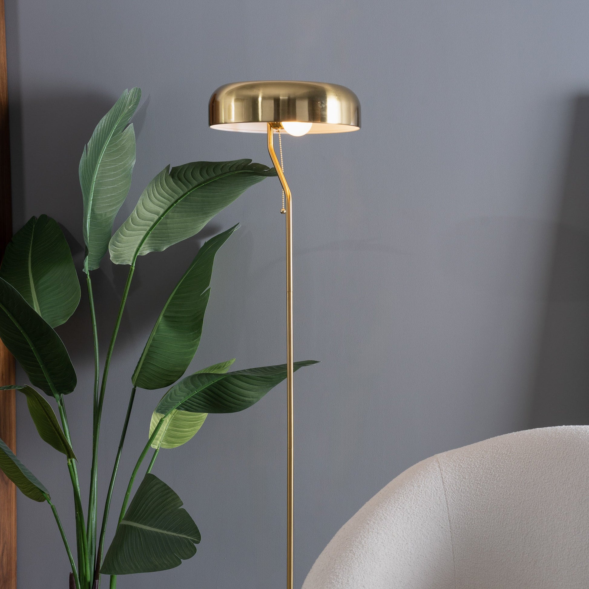 Gold floor lamp against a gray wall with a plant and chair in the foreground