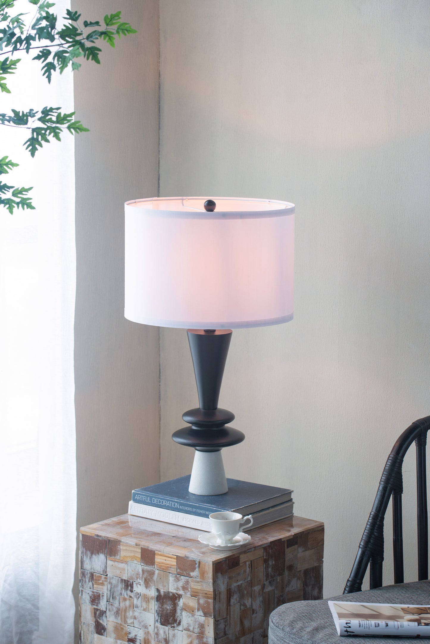 Table lamp with a white shade on a wooden side table next to a chair and plant.