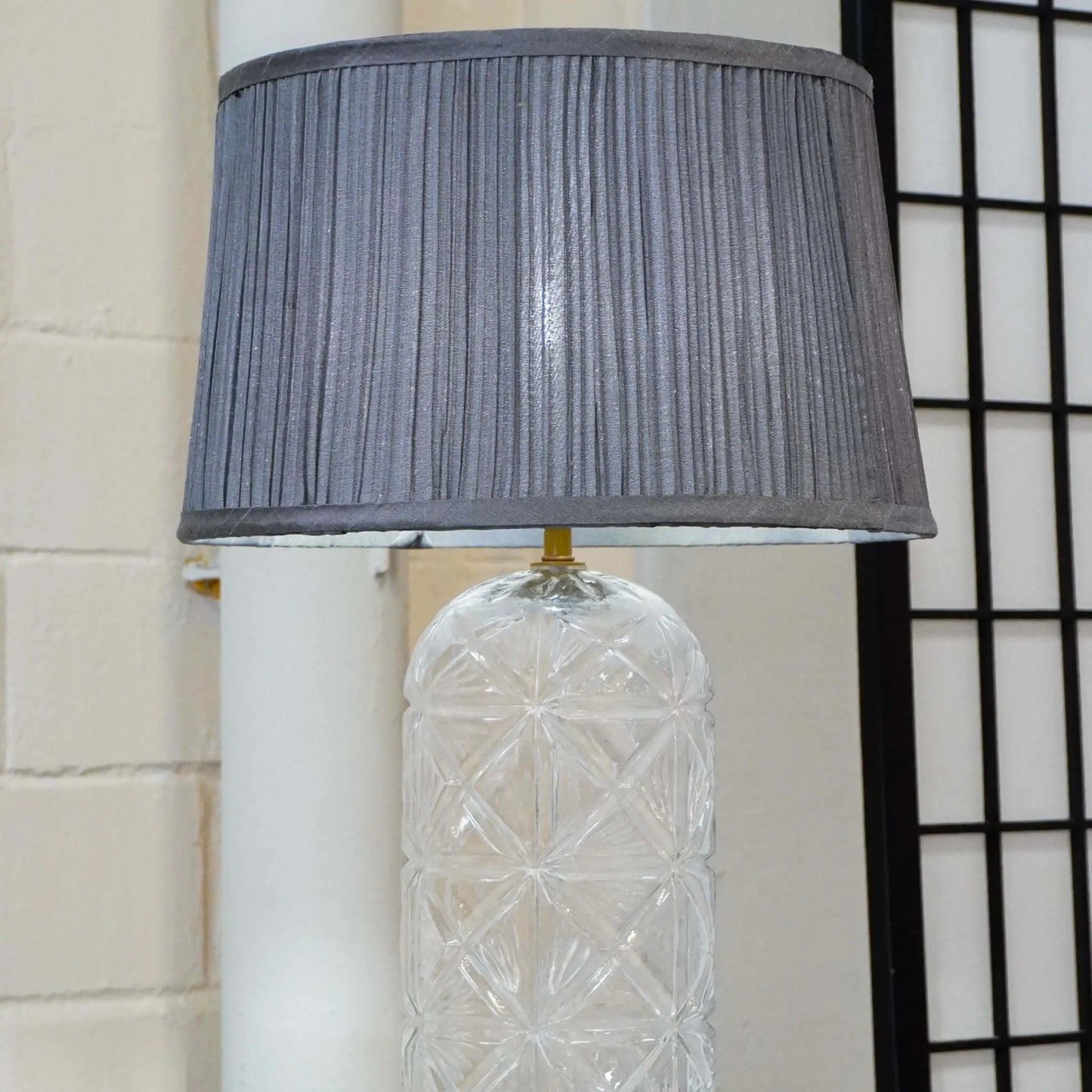 Decorative lamp with a textured clear base and gray pleated shade against a neutral wall.