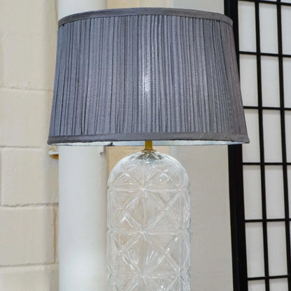Decorative lamp with a textured clear base and gray pleated shade against a neutral wall.