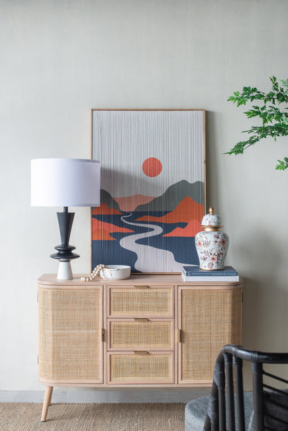 Table lamp on a wicker sideboard with decorative items including a vase and abstract art on a neutral wall.