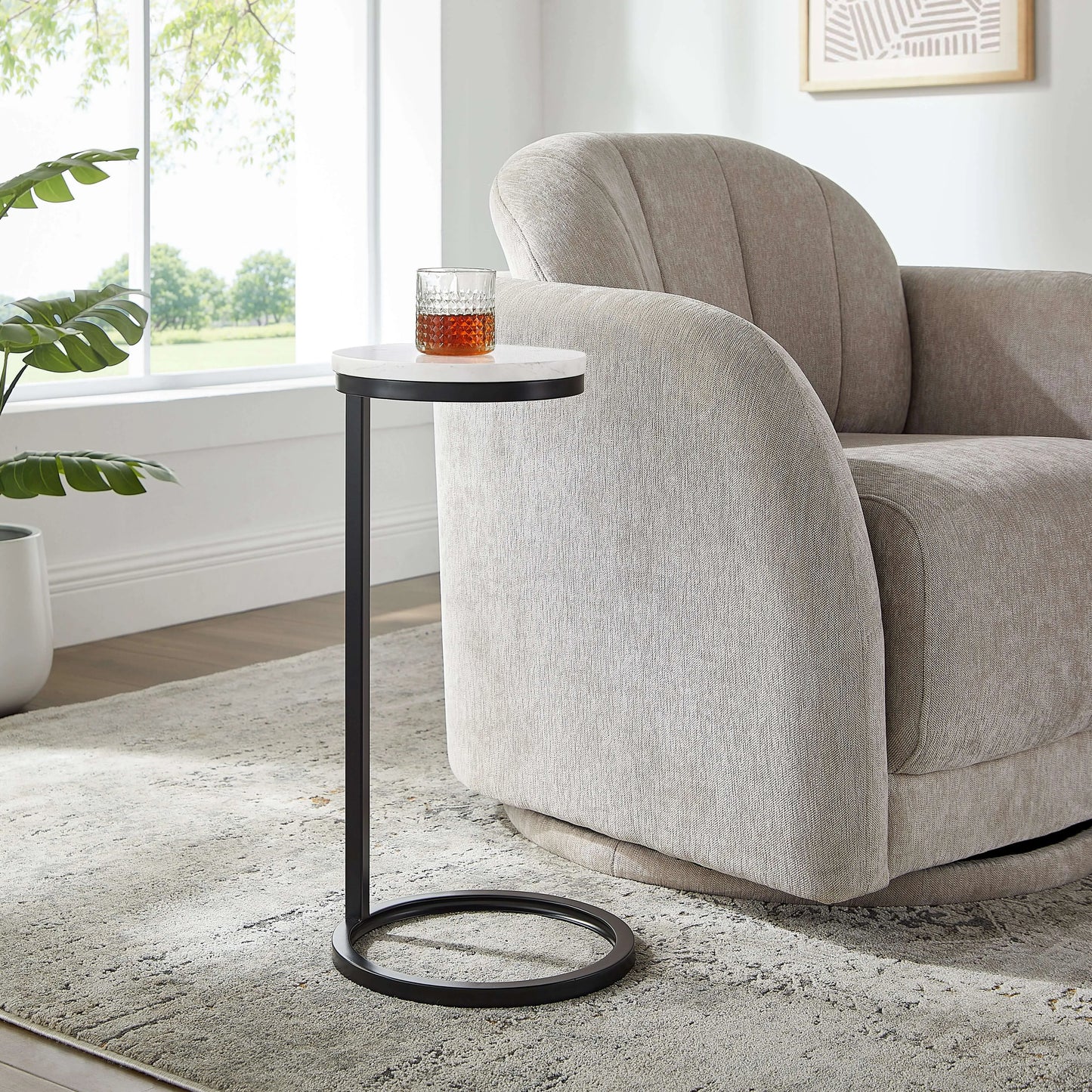 Small antique bronze end table with a glass of amber liquid next to a beige armchair in a room with a window and plant.