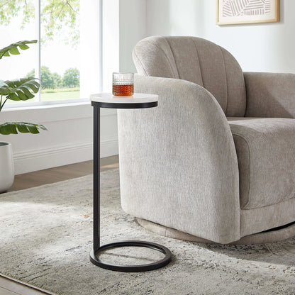 Small antique bronze end table with a glass of amber liquid next to a beige armchair in a room with a window and plant.