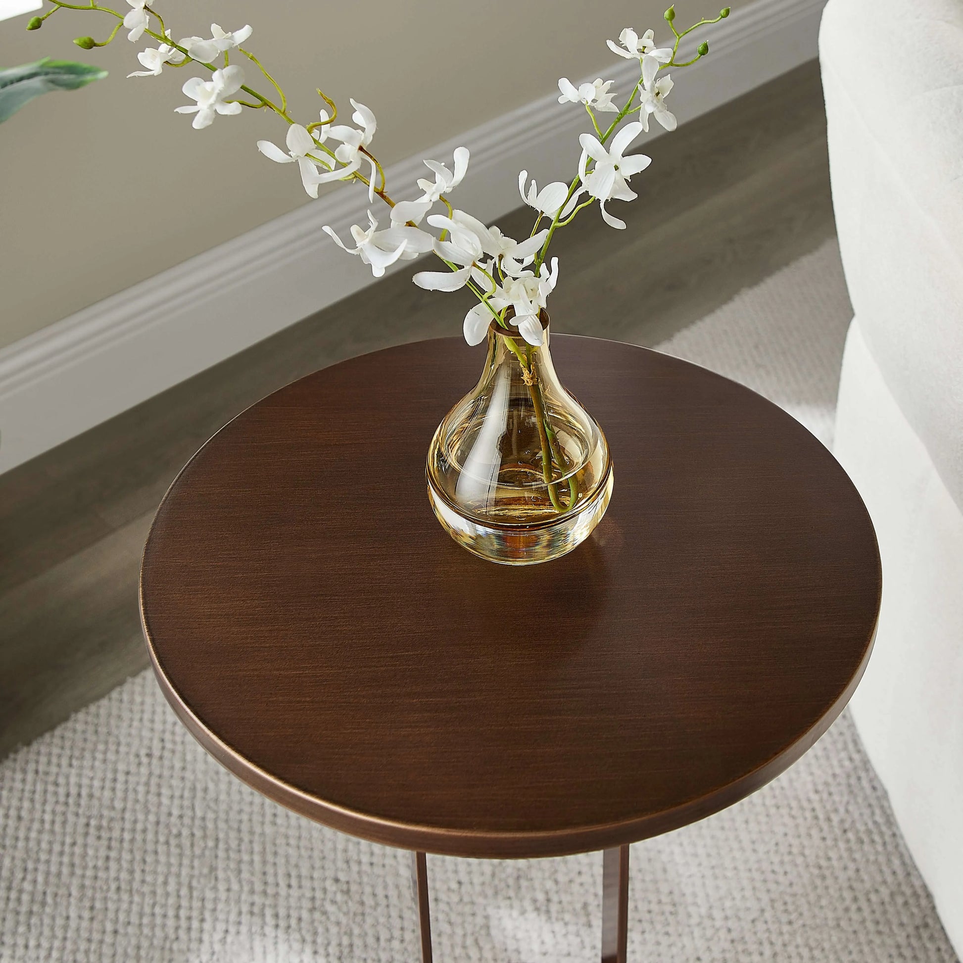 Round bronze end table with a clear vase containing white flowers on a neutral background
