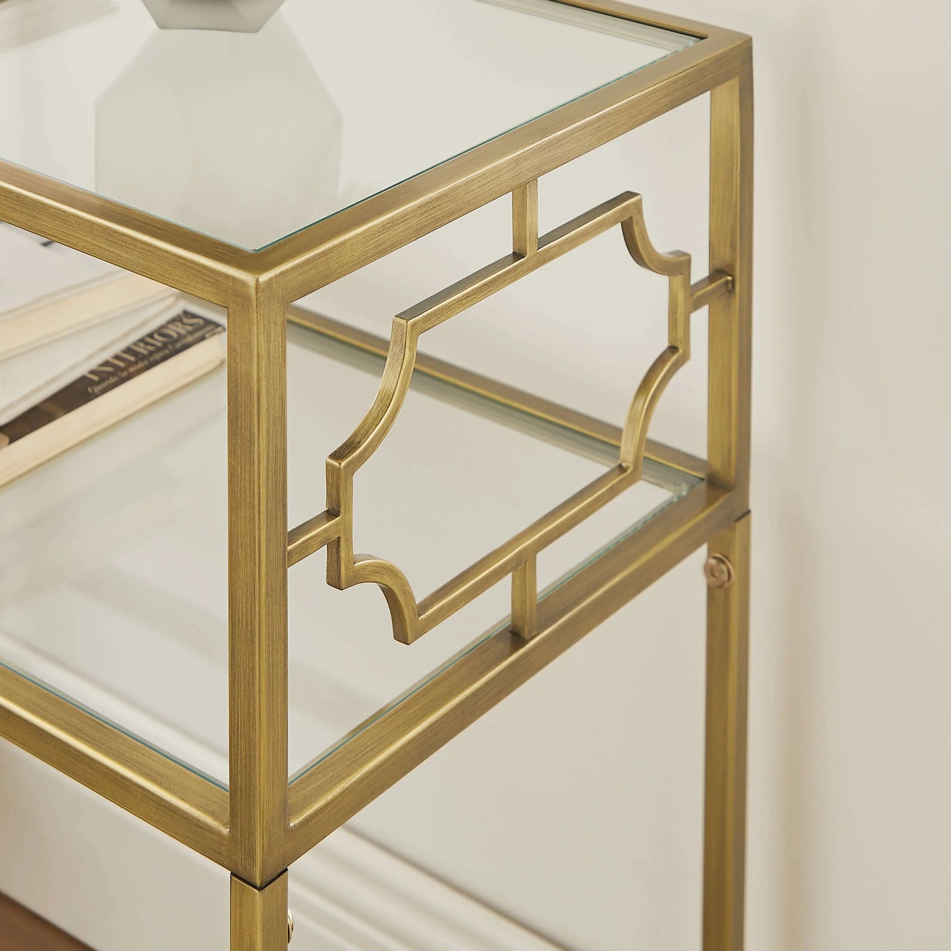 Gold-framed glass side table with 2 shelves and a white vase on a neutral background