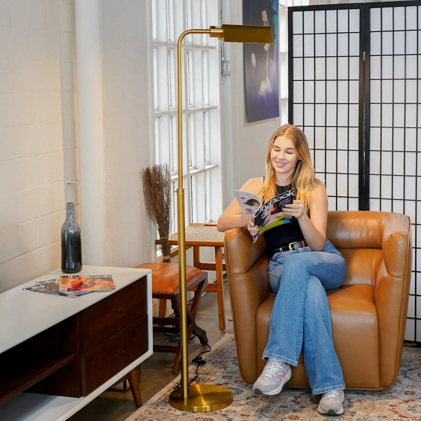Woman reading a magazine in a cozy living room with a brass floor lamp and wooden furniture.