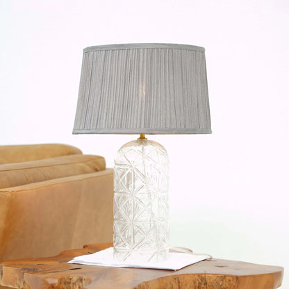 Table lamp with textured glass base and gray pleated shade on a wooden surface.