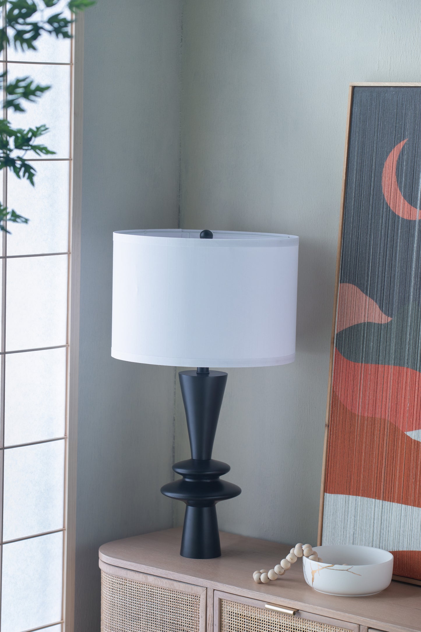 Table lamp with a white shade on a wooden side table next to a decorative painting.