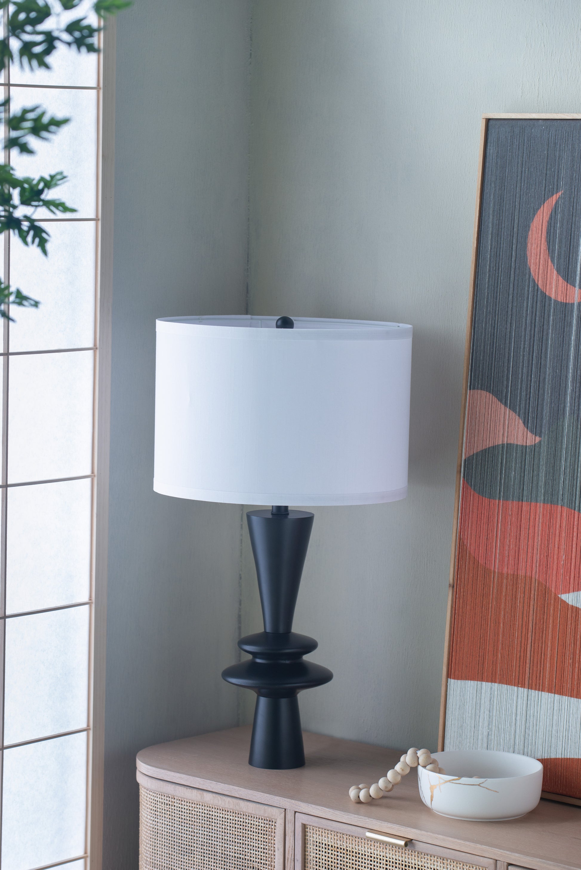 Table lamp with a white shade on a wooden side table next to a decorative painting.