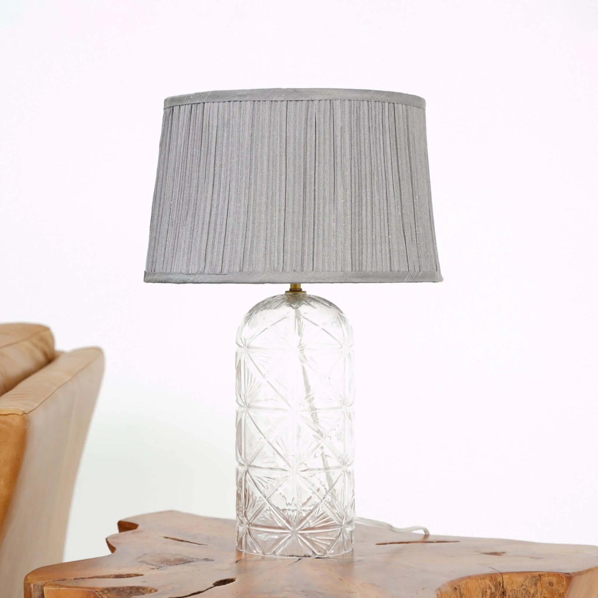 Table lamp with a clear glass base and gray pleated shade on a wooden surface.