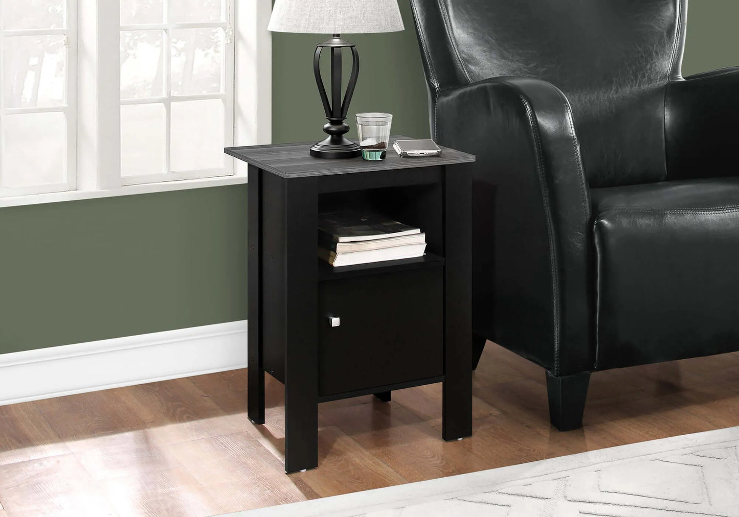 Black and grey end table with a lamp and books next to a black leather chair in a room with a window and green wall.