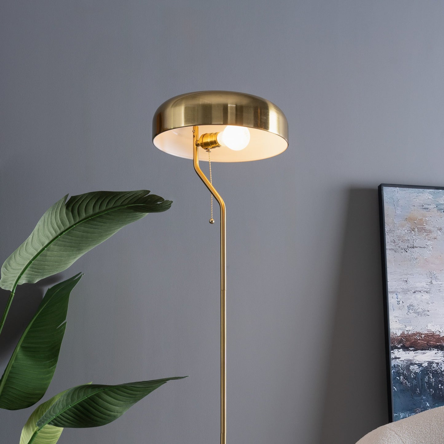 Gold floor lamp against a gray wall with a plant and painting in the background