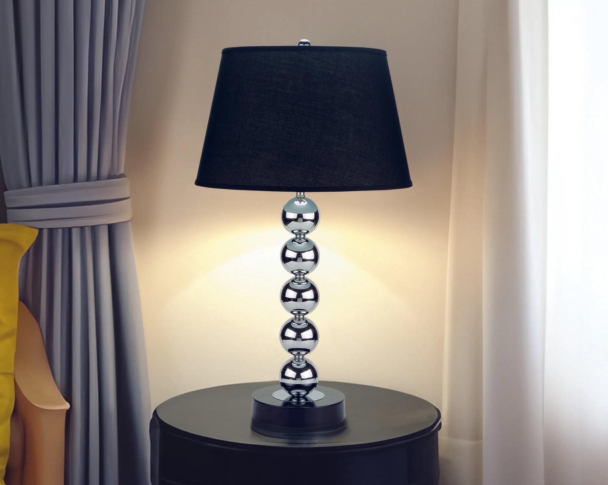 Table lamp with black shade and silver base on a round table against a curtain backdrop