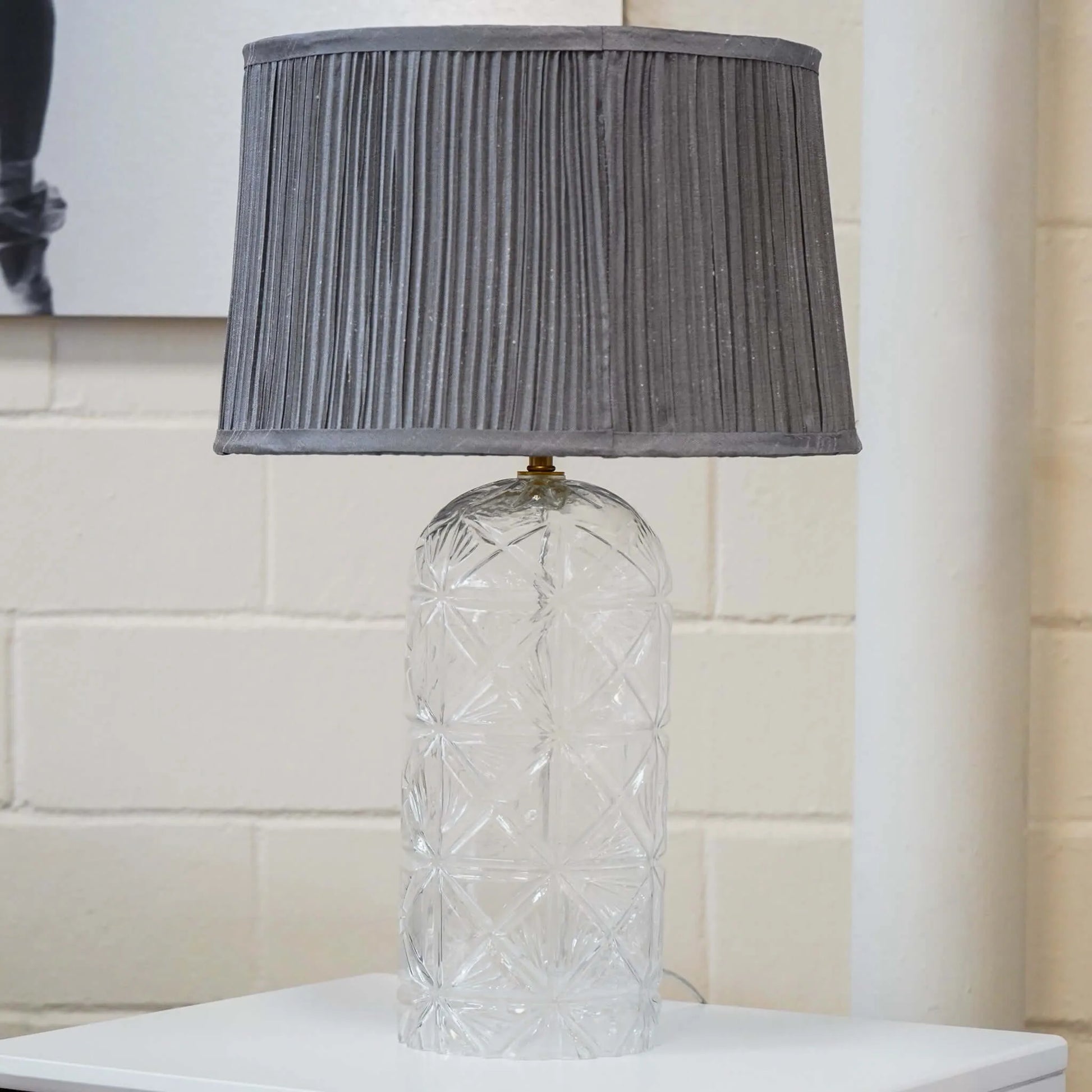 Table lamp with textured gray shade and clear glass base on a brick white surface.