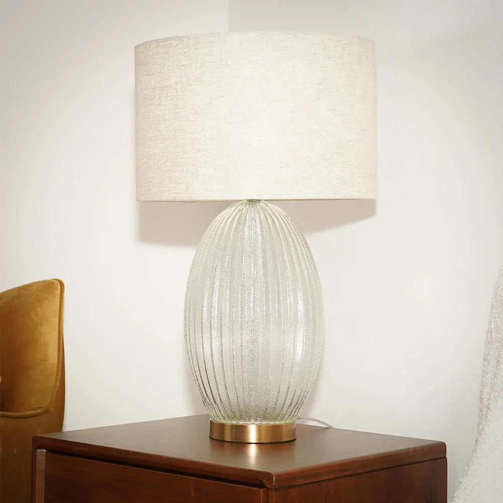Table lamp with ribbed glass base and beige shade on a wooden surface on a white background.