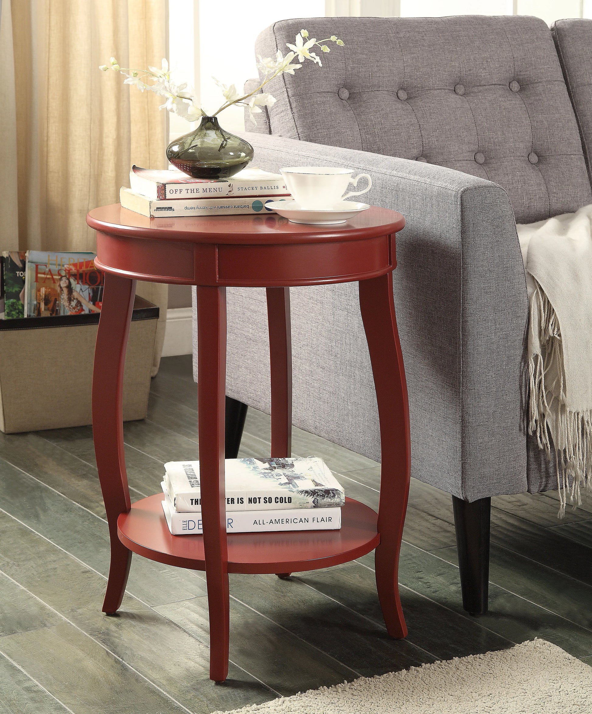 Red end table with books and a vase in front of a gray sofa.