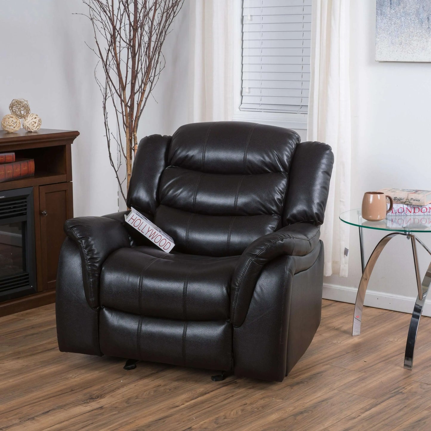 Black faux leather glider recliner chair in upright position in a living room setting with a fireplace and table.