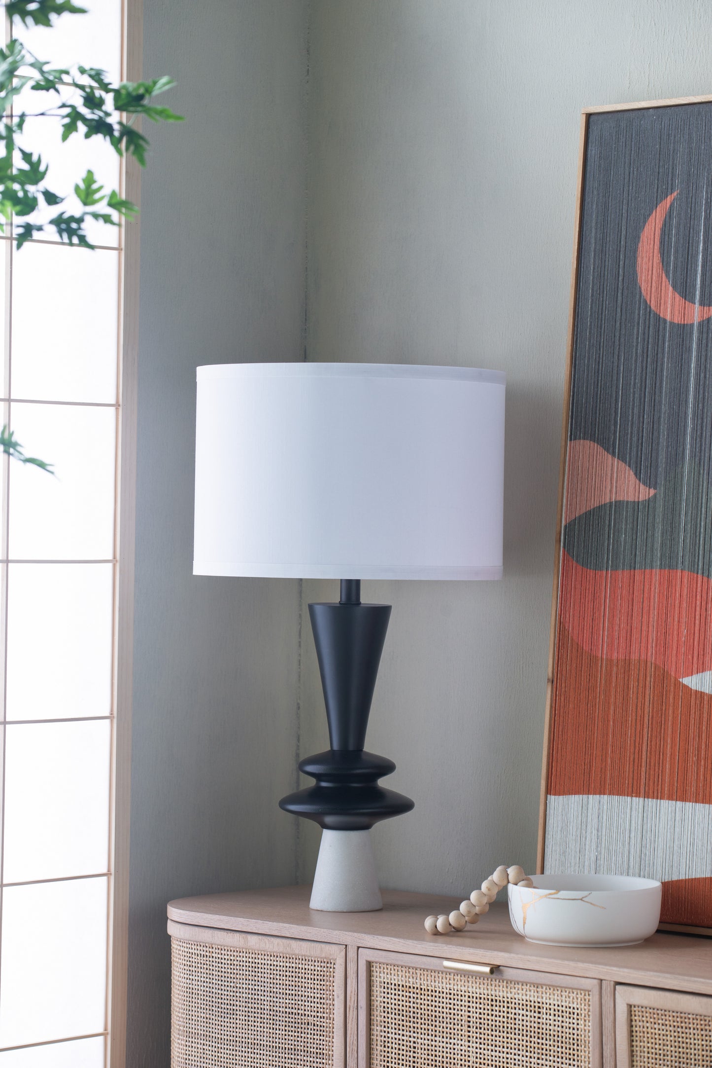 Table lamp with a white shade on a wooden cabinet against a wall with abstract art.