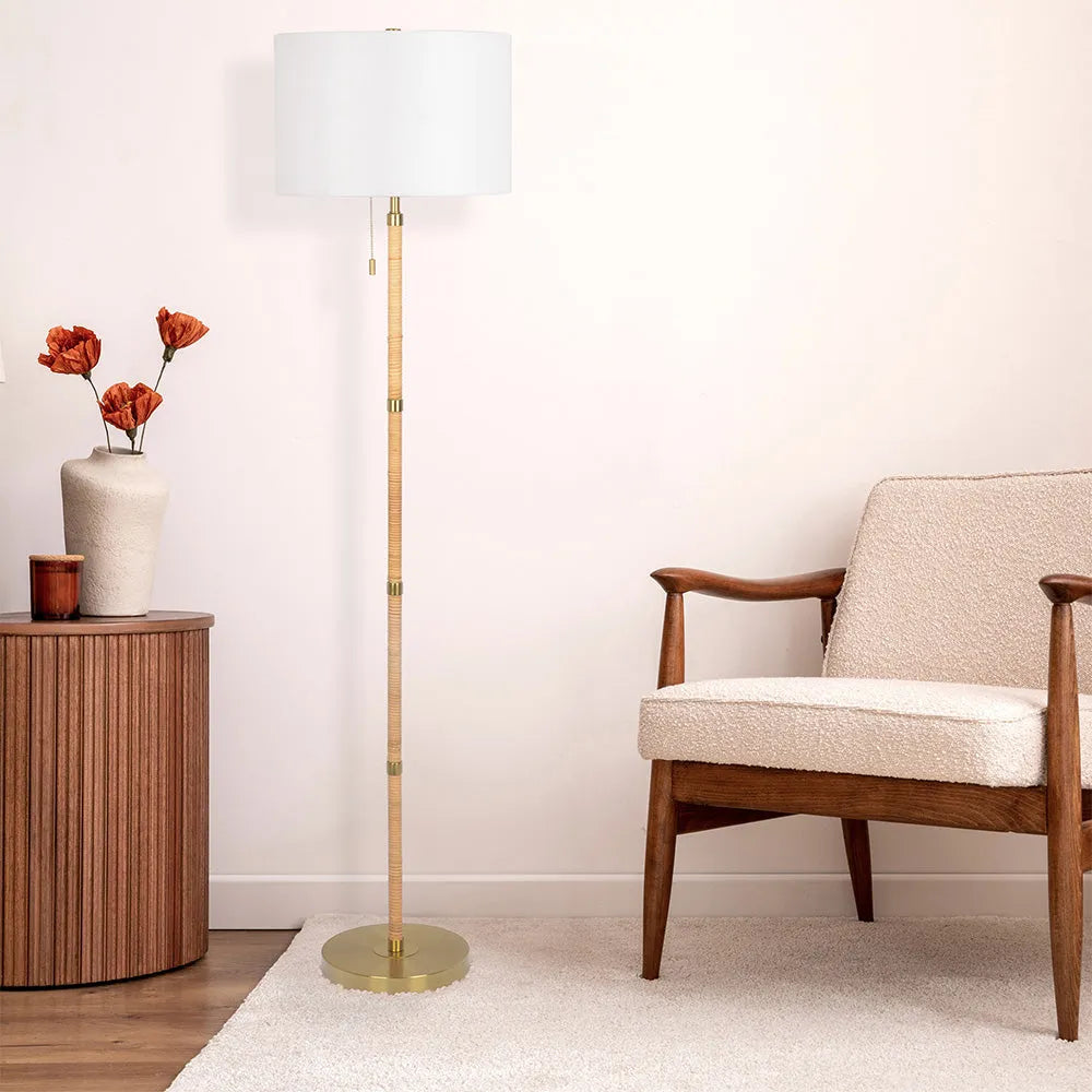 Floor lamp with a white shade next to a wooden side table with flowers and a chair in a room.