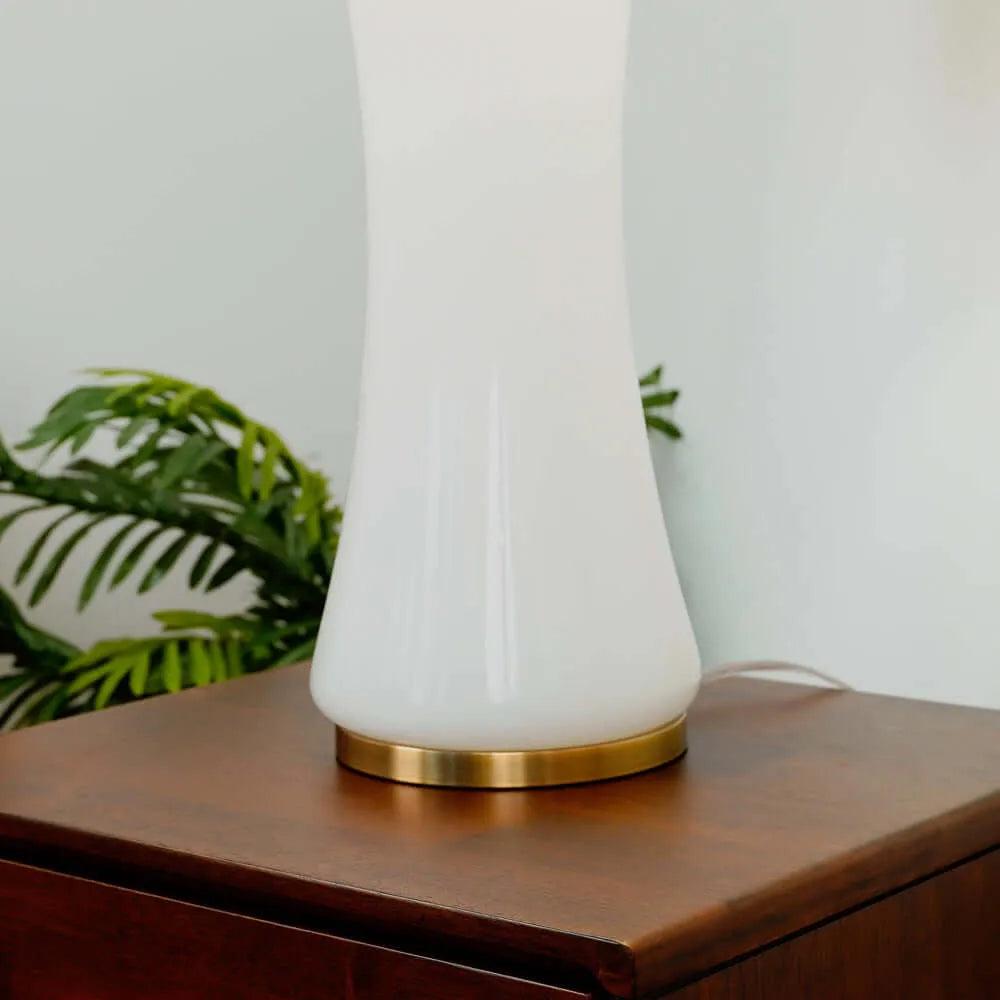 White table lamp with gold base on a wooden surface, blurred green plant in the background