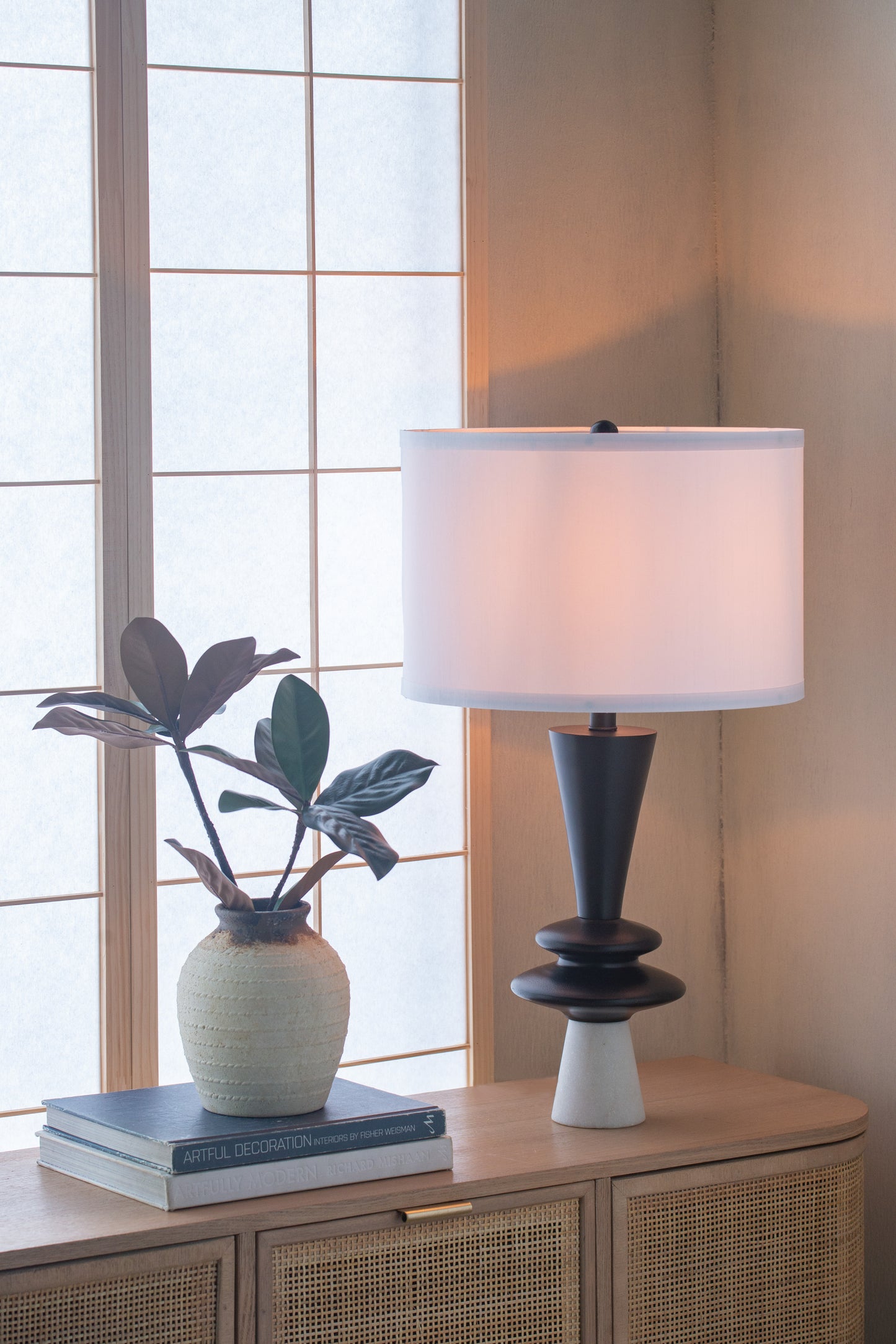 Table lamp with white shade on a wooden cabinet next to a plant and books, with a window in the background.