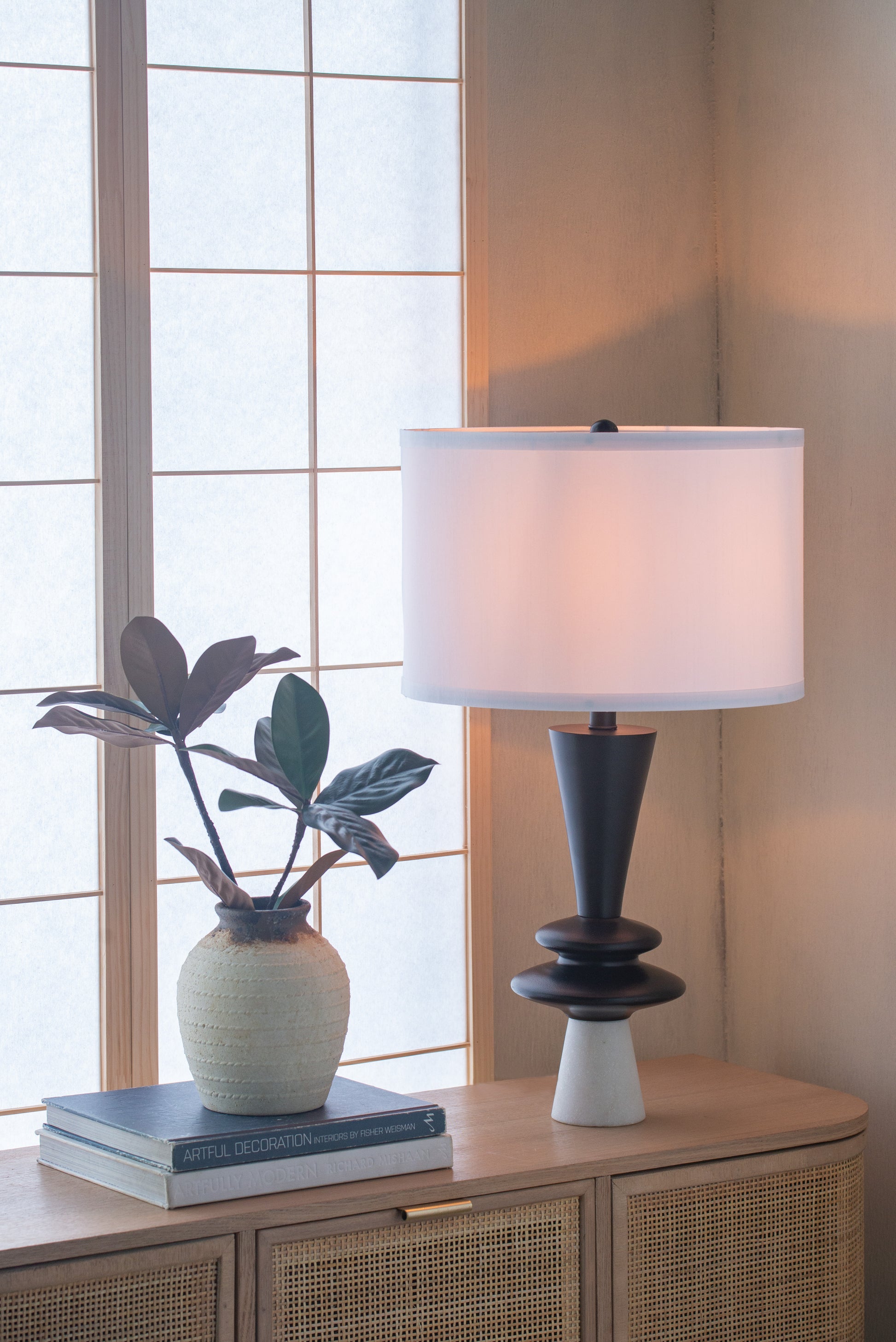 Table lamp with white shade on a wooden cabinet next to a plant and books, with a window in the background.