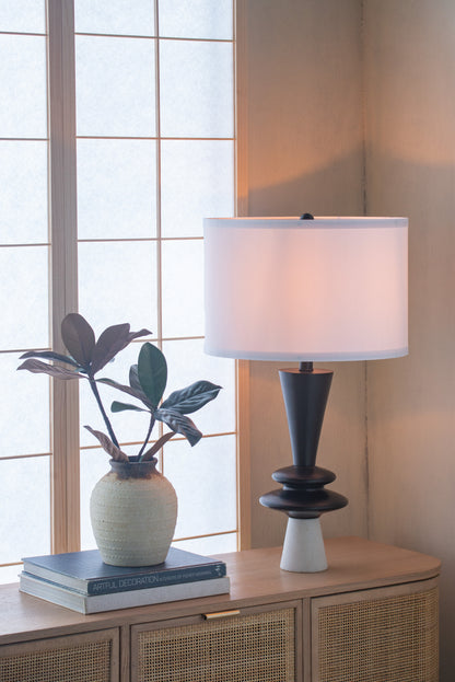 Table lamp with white shade on a wooden cabinet next to a plant and books, with a window in the background.