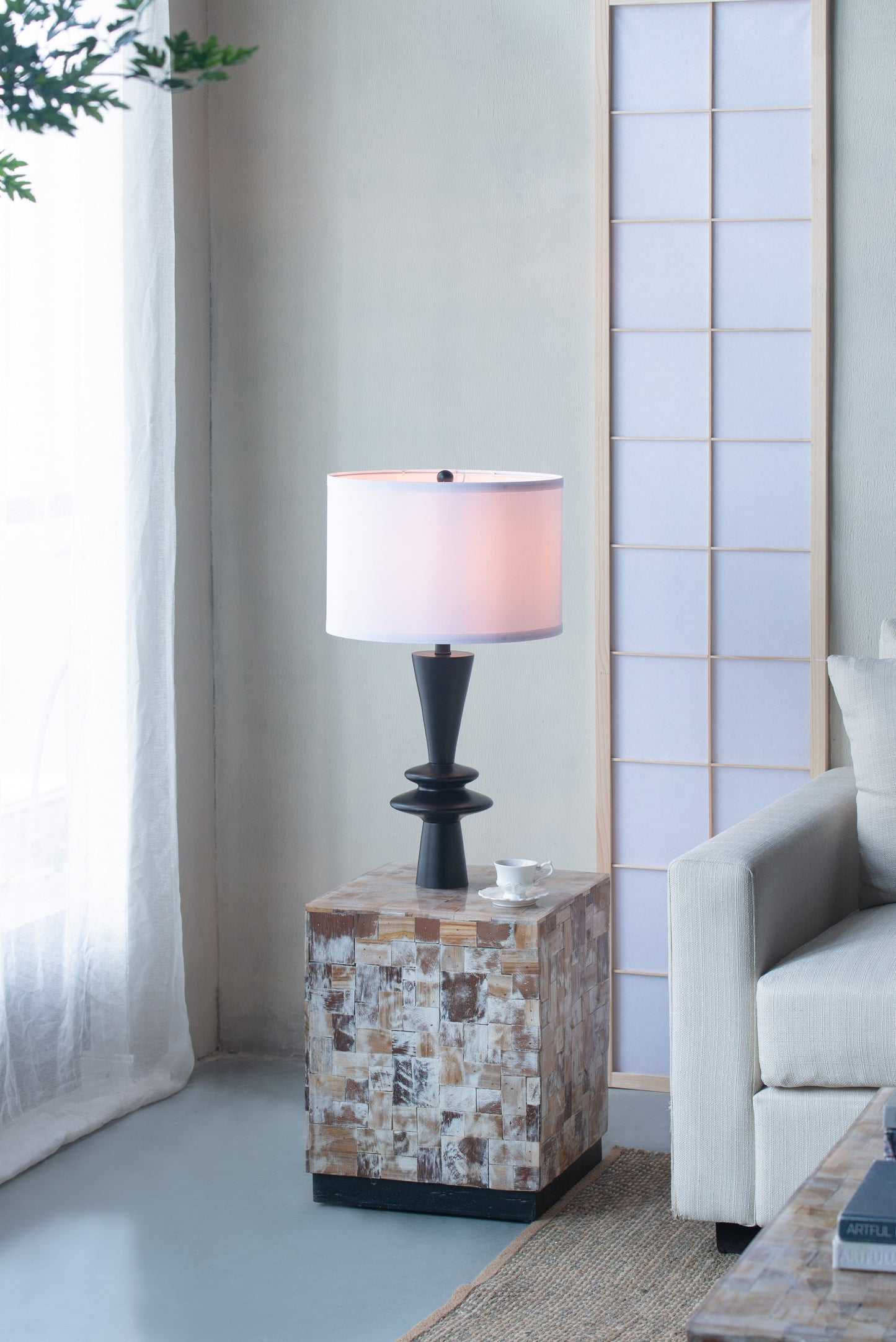 Table lamp on a stone pedestal in a living room setting with a sofa and window.